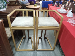 A Pair of bar stools feature a distinct bending frame design paired with soft, light-colored cushioned seats. W.47 D.50 H.90 Cm.